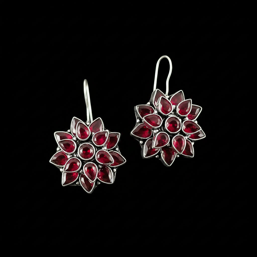 A pair of silver-toned dangle earrings with a starburst design set with deep red teardrop-shaped crystals, displayed on a dark gray reflective surface with the brand logo 'SAHELI MY BEST FRIEND' at the top.