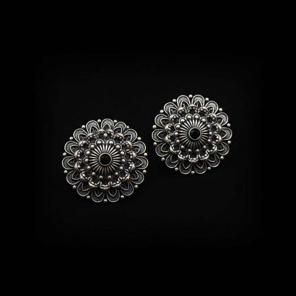 A pair of round, ornate silver-toned floral stud earrings with black oxidized detailing, displayed on a clean white background with the brand logo 'SAHELI MY BEST FRIEND' above them.