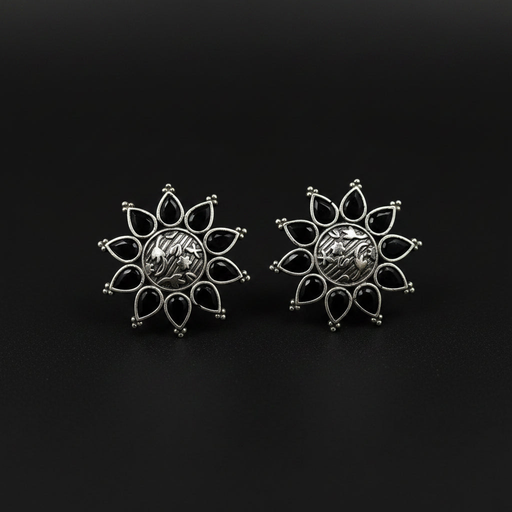A pair of silver-toned flower-shaped earrings with black teardrop inlays, displayed on a textured blue surface with a small white price tag in front. The brand name 'AL-RAWNAQ' is partially visible in gold lettering in the upper right corner.