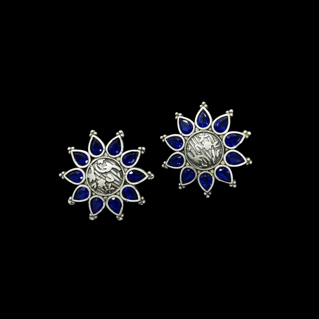 Two silver-toned floral stud earrings with deep blue inlay, displayed on a textured blue Al-Rawnaq box with a price tag held by a finger.