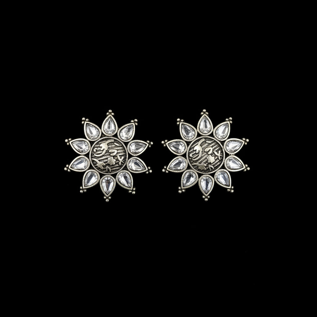 A pair of silver-toned crescent moon stud earrings with clear teardrop rhinestones, resting on a textured blue surface with the Al-Rawnaq brand logo visible at the top.