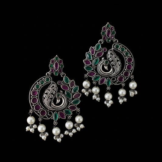 Ornate silver earrings with green, purple, and pearl accents on a dark background