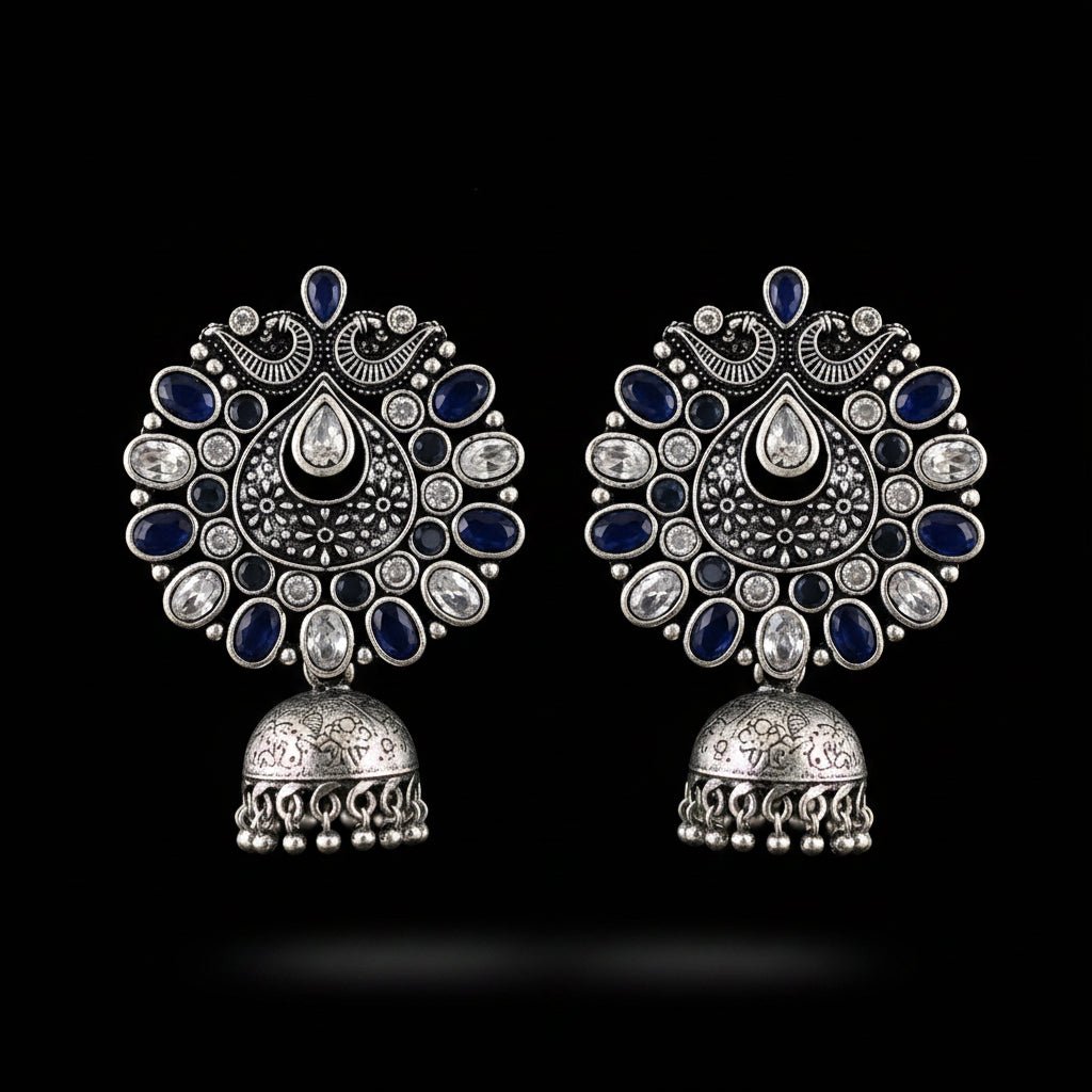 A pair of ornate silver-toned Indian jhumka earrings decorated with dark blue enamel and clear rhinestones, displayed on a black jewelry stand.