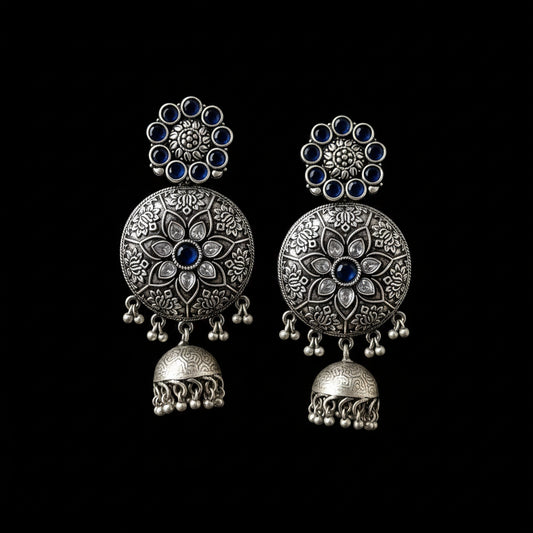 Silver floral earrings with blue stones on a gray stand, branded 'SAHELI'.
