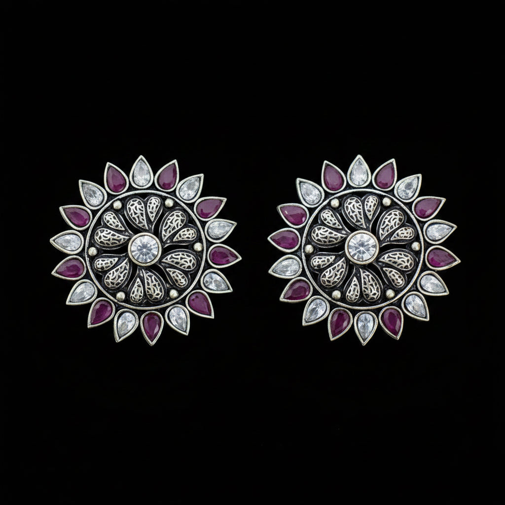 A pair of silver-toned floral stud earrings with purple gemstone petals and clear central stones, displayed on a dark gray reflective surface.
