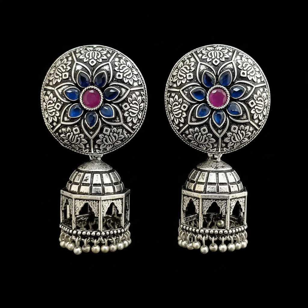 A pair of ornate silver-toned Indian jhumka earrings displayed on a gray Y-shaped jewelry stand. The earrings feature circular studs with intricate floral filigree, blue enamel accents, and a central pink stone, hanging from silver bell-shaped dangles with small pearl-like beads at the bottom.
