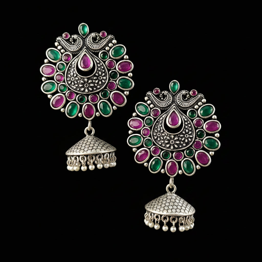 A pair of ornate Indian silver-toned jhumka earrings with green and pink stones, displayed on a gray stand against a neutral gray background with the brand name 'SAHELI MY BEST FRIEND' at the top.