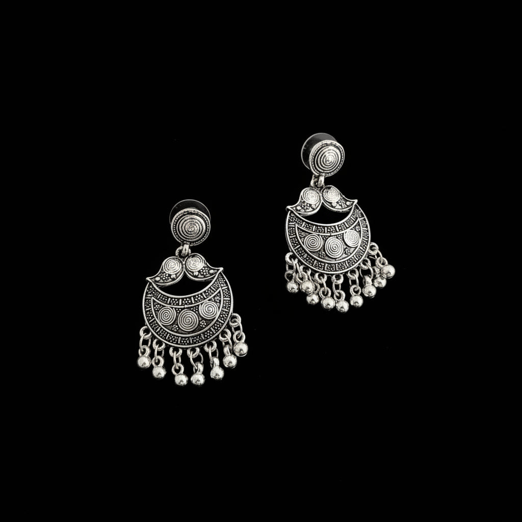 A pair of silver-toned traditional Indian jhumka earrings with intricate filigree work and dangling bead chains, displayed on a dark reflective surface.