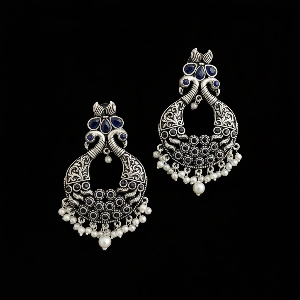 A pair of silver-toned traditional Indian jhumka earrings with blue stone accents and white bead fringe, displayed on a dark reflective surface.