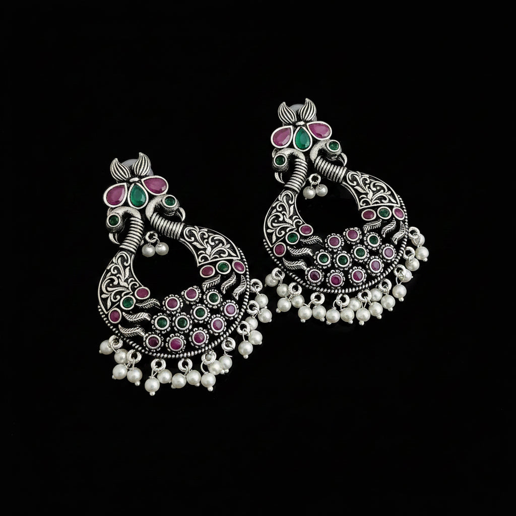 A pair of ornate silver-toned Indian jhumka earrings decorated with pink, green, and white stones, displayed on a dark reflective surface.