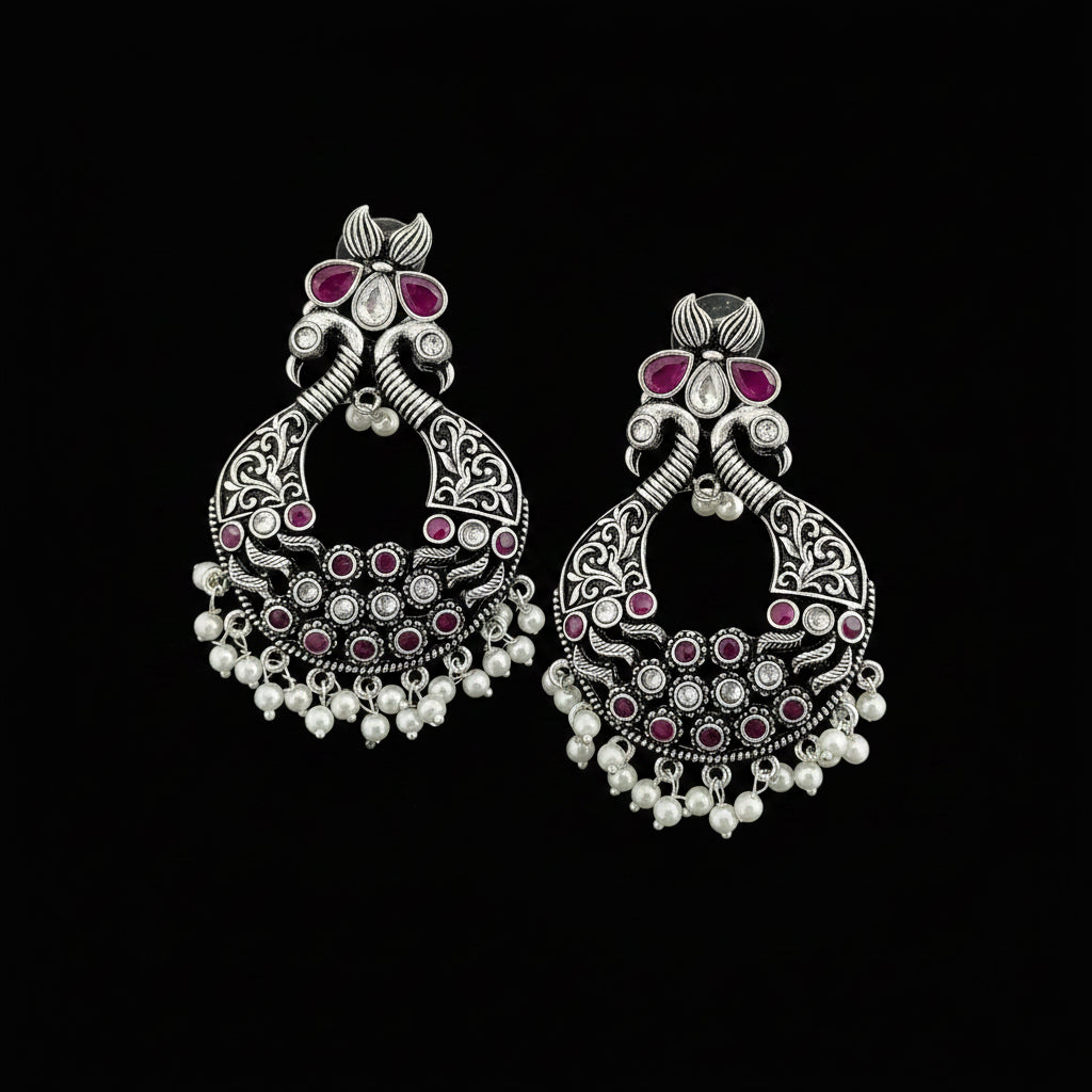 A pair of ornate silver-toned Indian jhumka earrings decorated with pink stones and white bead fringe, displayed on a dark reflective surface.