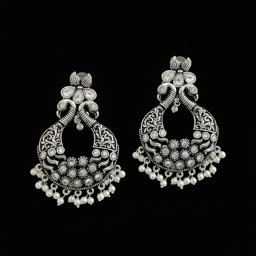 A pair of ornate silver-toned traditional Indian jhumka earrings decorated with intricate filigree work, small clear stones, and dangling white pearl beads, displayed on a dark reflective surface.