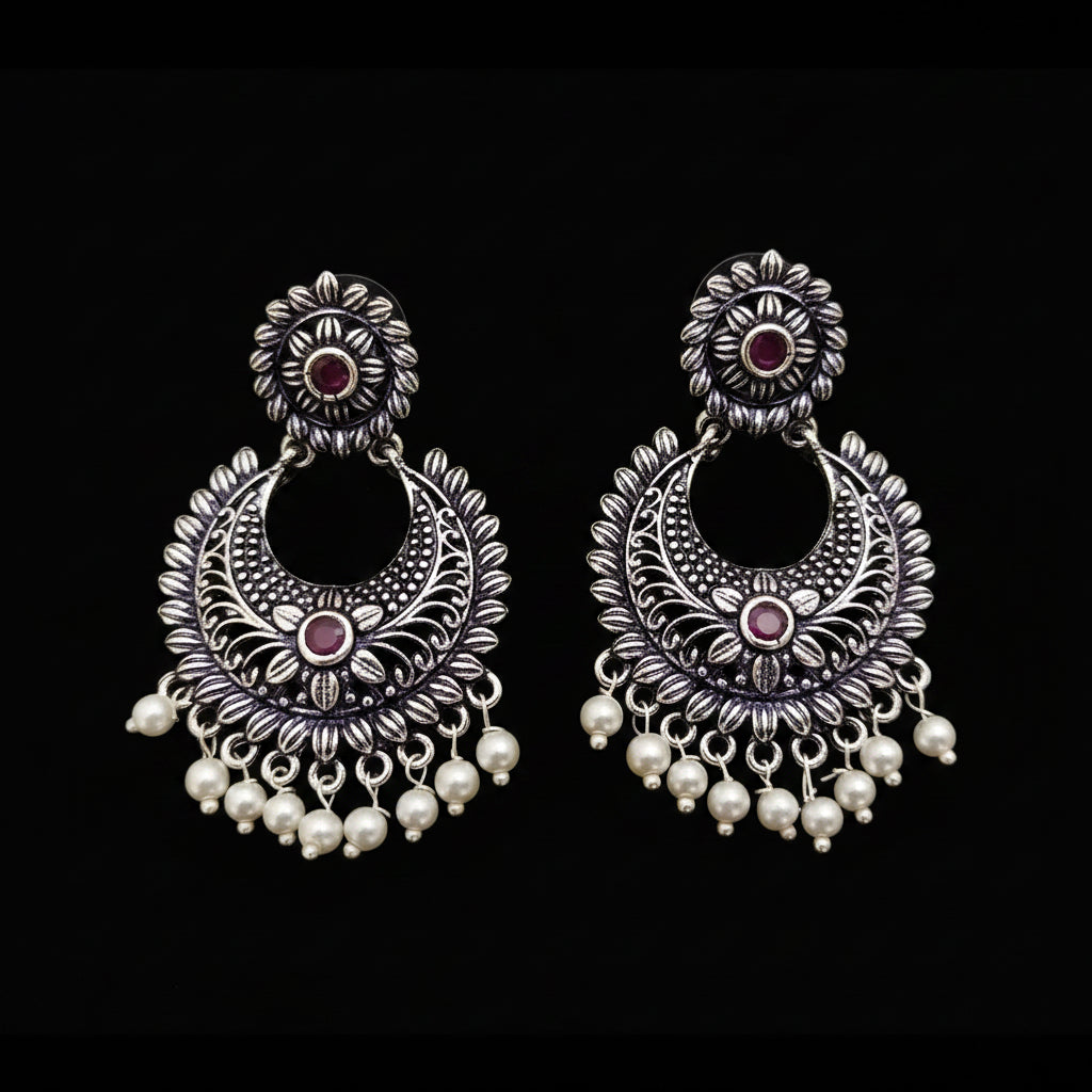 Pair of silver earrings with red stones and pearl drops on a gray background, branded 'Saheli'.
