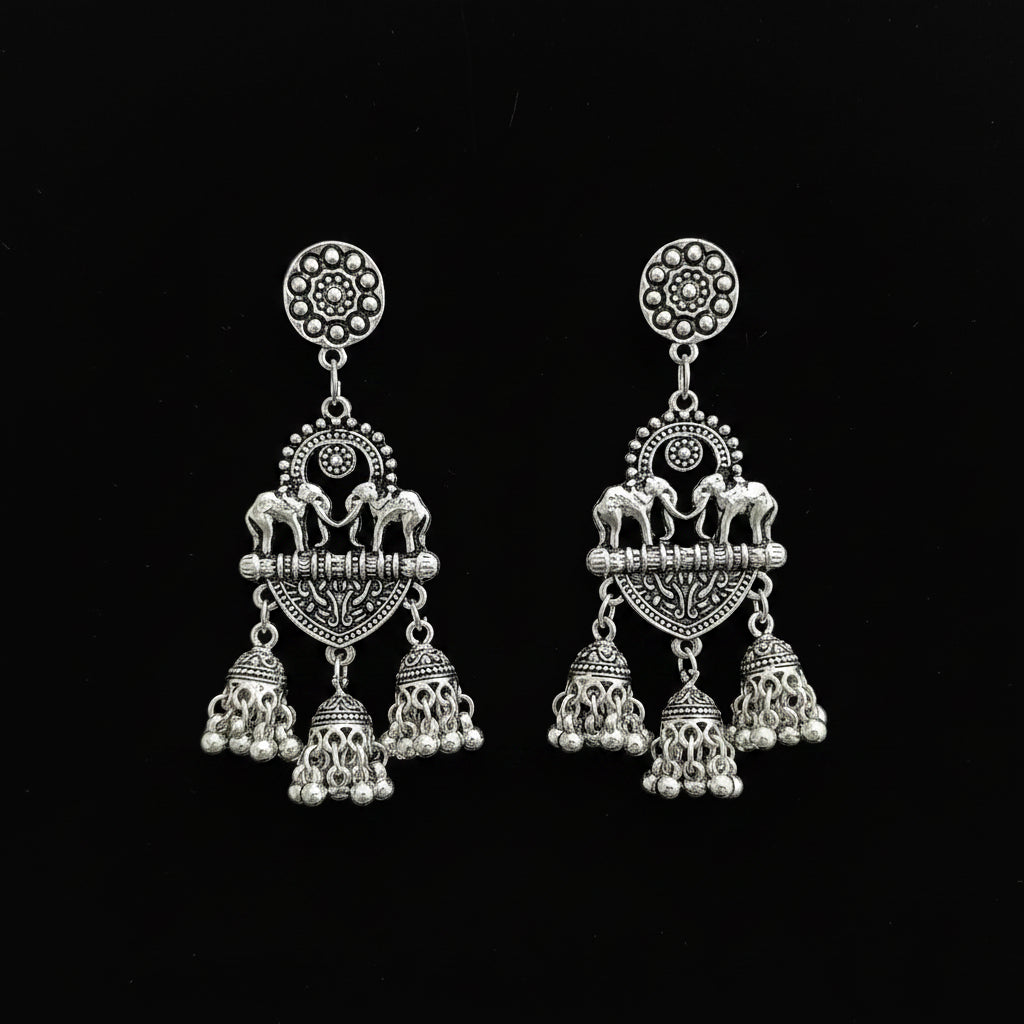 A pair of silver-toned Indian-style dangle earrings with elephant and floral motifs, displayed on a black stand against a dark reflective surface.