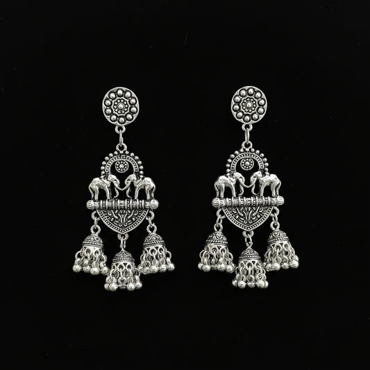 A pair of silver-toned Indian-style dangle earrings with elephant and floral motifs, displayed on a black stand against a dark reflective surface.