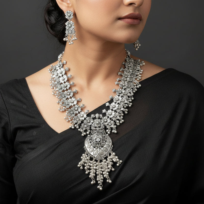 Woman wearing a silver necklace and earrings with a dark background, featuring the brand 'Sahil'.