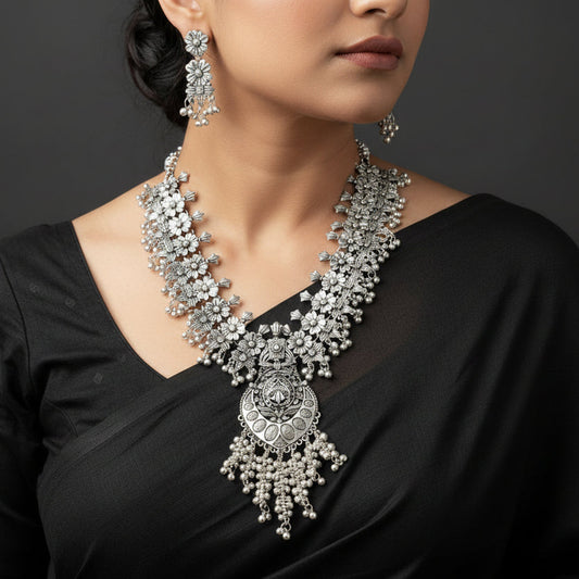 Woman wearing a silver necklace and earrings with a dark background, featuring the brand 'Sahil'.