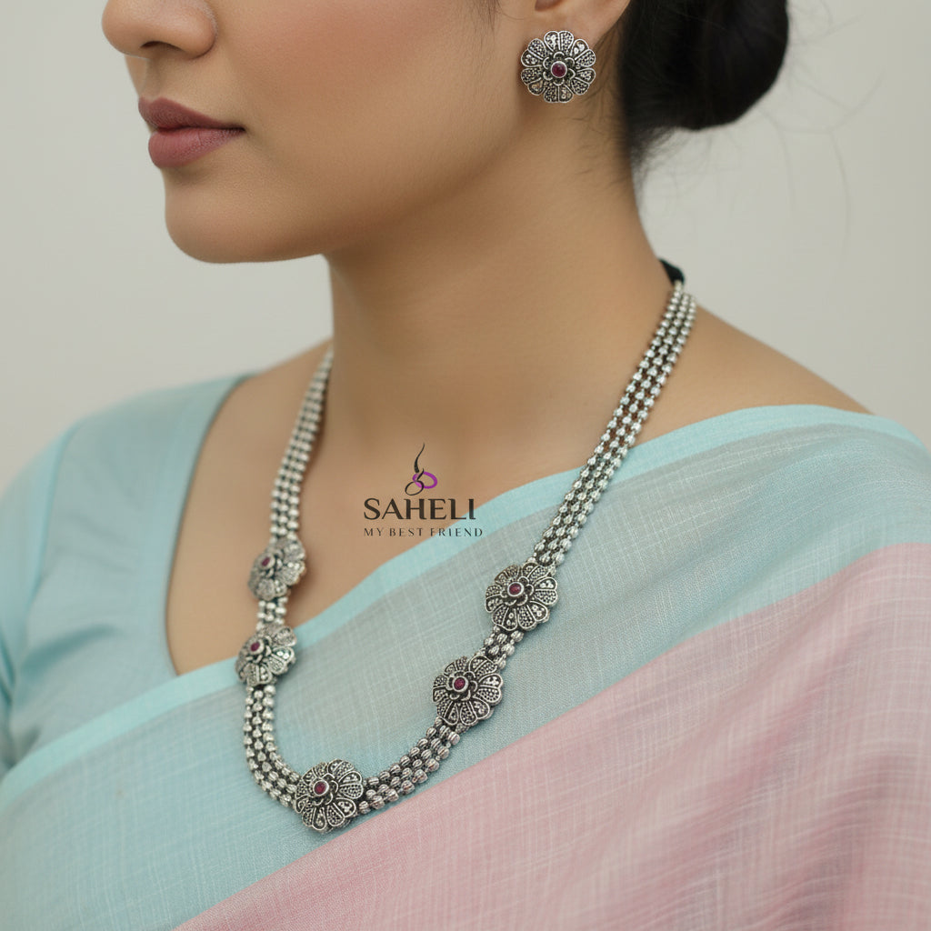 Silver necklace with floral designs and red stones on a dark background, branded 'Saheli'.