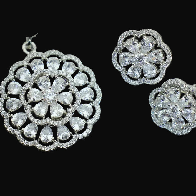 Silver floral earrings with clear stones on a reflective surface, branded 'Saheli'.