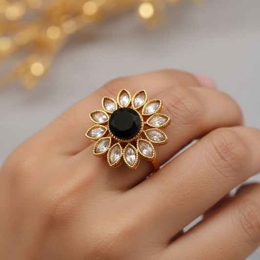 A gold-toned ring shaped like a lotus flower, featuring a central black oval stone surrounded by small pale pink stones, shown on a gray background with a white product tag attached.