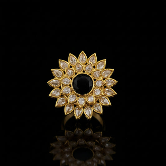 A gold-toned floral brooch featuring a central black oval stone surrounded by clear teardrop-shaped stones, displayed on a dark reflective surface with the SAHELI brand logo in the top left corner.
