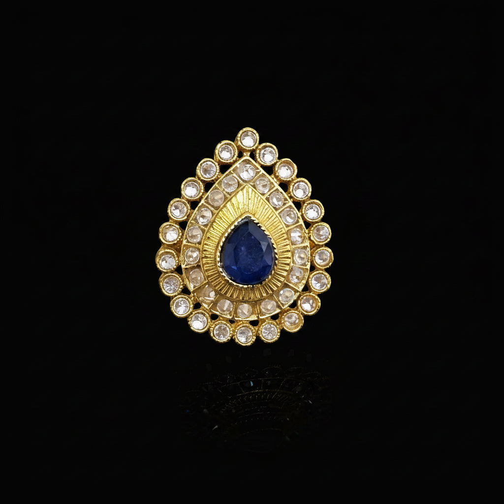 A gold teardrop-shaped ring with a central deep blue teardrop gemstone and surrounding clear rhinestones, displayed on a reflective gray surface. The brand logo 'SAHELI MY BEST FRIEND' is visible in the top left corner.