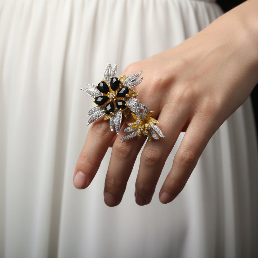 Gold and silver flower-shaped jewelry piece with black stones on a gray background, featuring the brand 'SAHELI'.