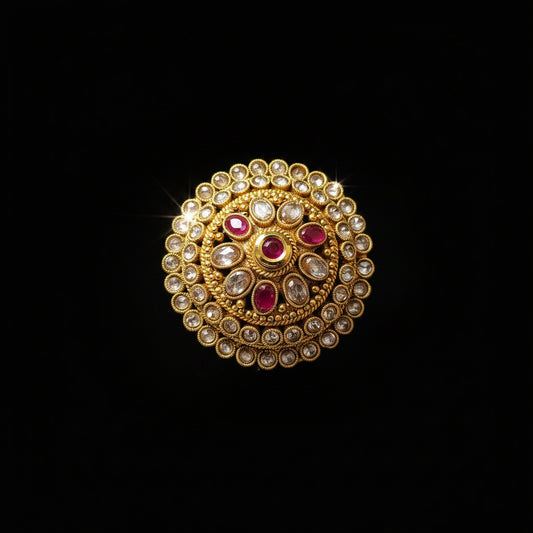 A gold-toned Indian-style bridal ring with a circular mandarin design, featuring clear stones in outer rings, a central gold floral motif, and three pink gemstones, shown on a reflective gray surface with the SAHELI MY BEST FRIEND logo in the top left.