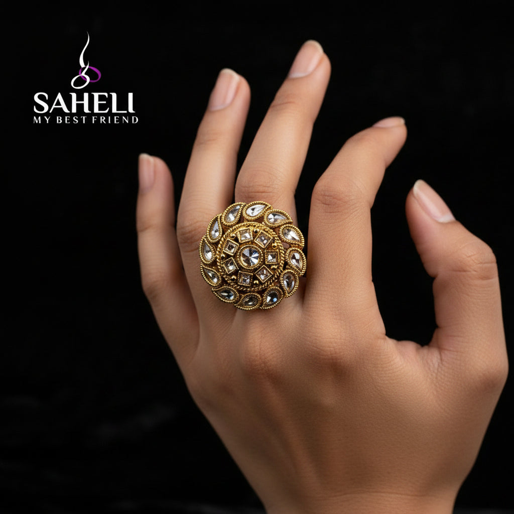 A large, ornate gold ring with intricate stone detailing is displayed on a black velvet glove hand against a dark background. The brand logo 'SAHELI MY BEST FRIEND' is overlaid near the top of the hand.