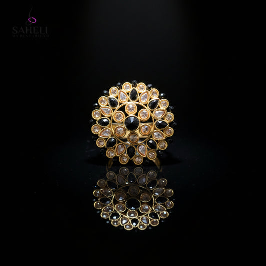 A gold-toned circular Kundan choker brooch featuring black stones and clear stones, displayed on a dark gray reflective surface with a Saheli My Best Friend logo in the top left corner.