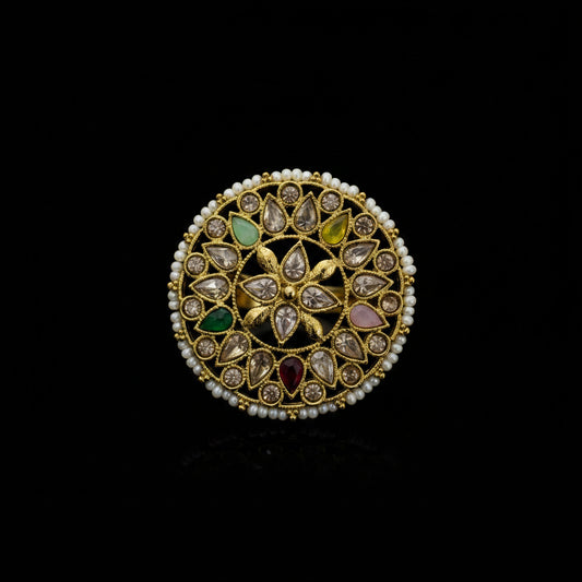 A gold-plated Indian Kundan ring with a circular filigree design, decorated with small white pearls, green, yellow, pink, and red stones, shown on a dark reflective surface.