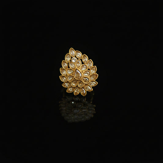 A gold ring featuring a detailed, scalloped floral or lotus-like design set with clear stones, displayed on a dark reflective surface.