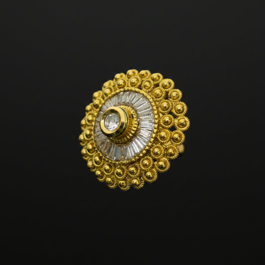 A gold-toned traditional Indian ring with a circular filigree design, a central clear stone, and a white inlay, shown on a dark reflective surface. The SAHELI brand logo is visible in the top left corner.