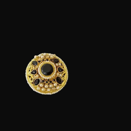 Gold ring with black stones on a black hand, branded 'SAHELI'.