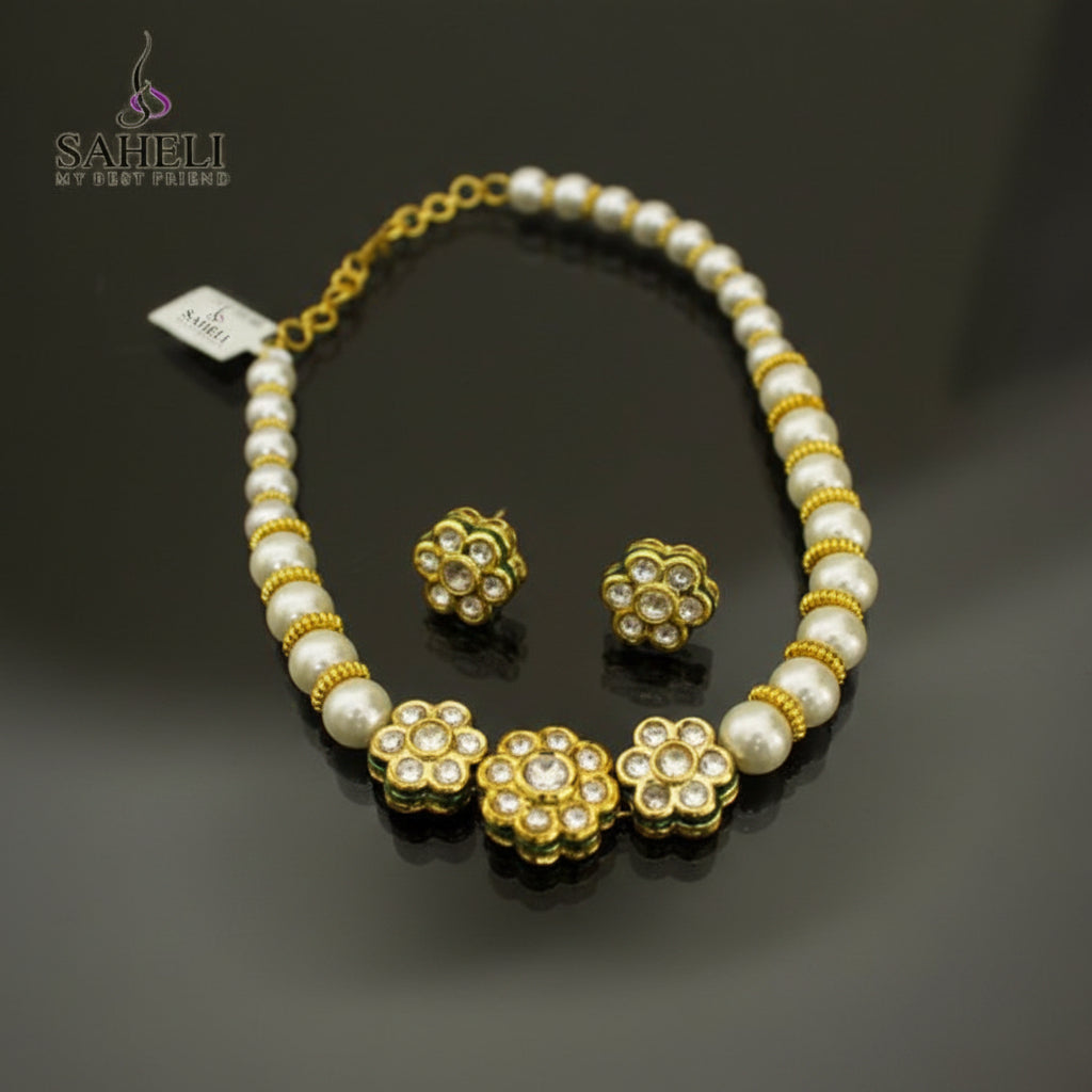 A gold and white beaded necklace with matching flower-shaped stud earrings, displayed on a dark reflective surface. The necklace features white pearl-like beads separated by gold spacer rings and three larger gold flower motifs with clear stones.