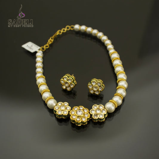 A gold and white beaded necklace with matching flower-shaped stud earrings, displayed on a dark reflective surface. The necklace features white pearl-like beads separated by gold spacer rings and three larger gold flower motifs with clear stones.