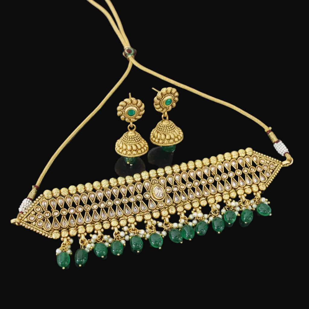 Gold-toned traditional jewelry set featuring a choker necklace with dangling green bead accents, matching gold earrings with green stones, displayed on a black reflective surface. The brand name 'SAHELL MY BEST FRIEND' is visible in the top left corner.