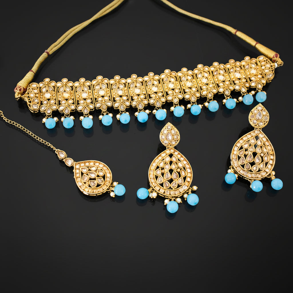 Gold-toned Indian bridal jewelry set featuring a choker necklace, matching earrings, and a maang tikka, embellished with white stones and light blue bead accents, displayed on a dark reflective surface with the SAHeli brand logo.