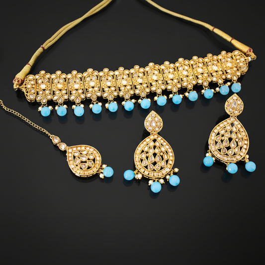 Gold-toned Indian bridal jewelry set featuring a choker necklace, matching earrings, and a maang tikka, embellished with white stones and light blue bead accents, displayed on a dark reflective surface with the SAHeli brand logo.
