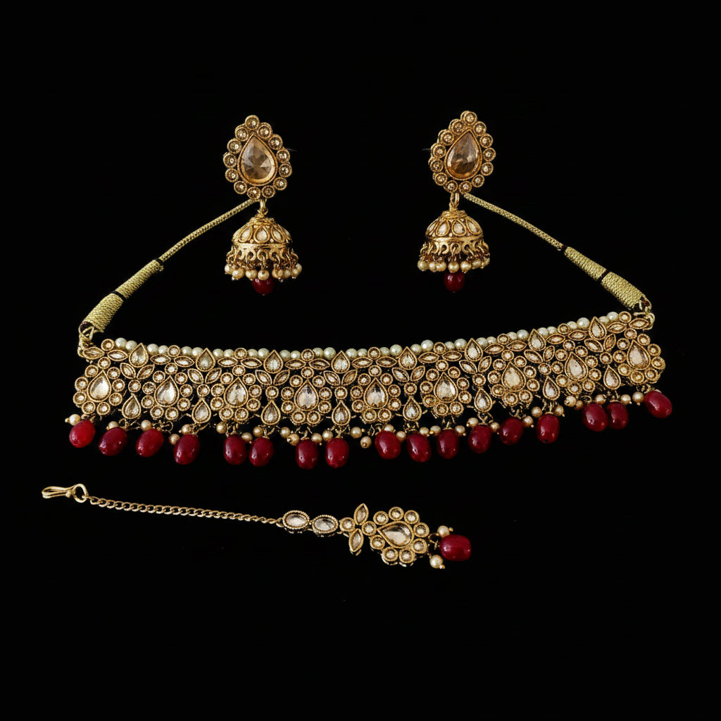 Gold-toned Indian bridal jewelry set including a choker necklace, matching earrings, and a maang tikka, decorated with cream stones and red beads, displayed on a black background. Saheli My Best Friend logo is visible in the top left.