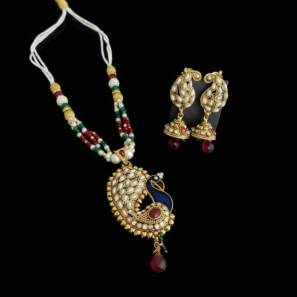 Gold-plated peacock pendant necklace and matching jhumka earrings with white, red, and green beaded accents, displayed on a dark reflective surface. The brand logo 'SAHELI MY JEWELLERY' is visible in the top right.