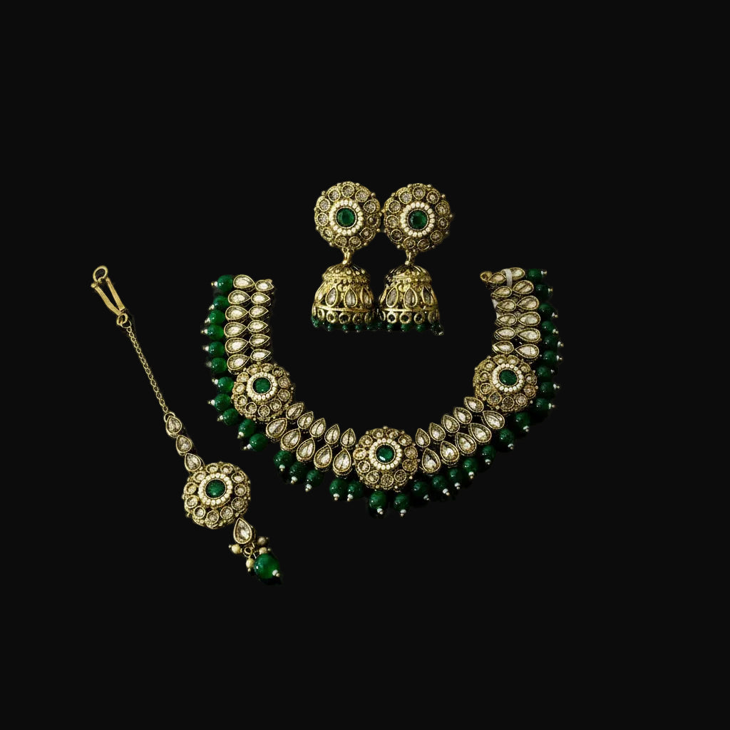 Gold and green traditional Indian jewelry set including a necklace, earrings, and a maang tikka, displayed on a black reflective surface with the Saheli brand logo in the top left corner.
