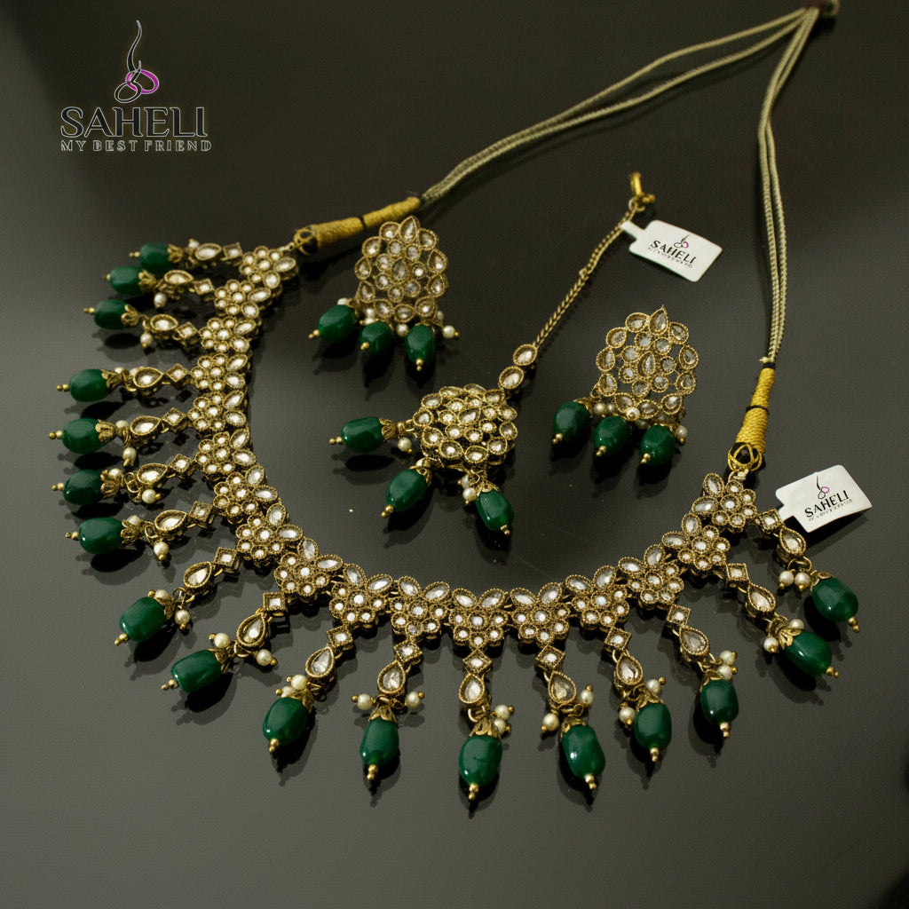 Gold-plated Kundan necklace and matching earrings with green stone accents, displayed on a dark reflective surface. The brand logo 'SAHELI MY BEST FRIEINDS' is visible in the top left, and a small white tag is attached to the necklace.