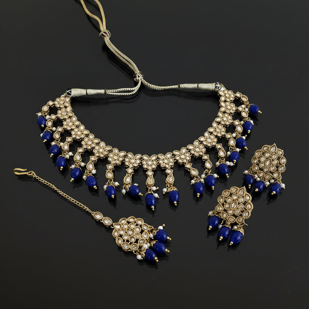Gold-plated Indian bridal jewelry set featuring a statement necklace, matching earrings, and a maang tikka, embellished with deep blue beads and clear stones, displayed on a dark reflective surface with the brand logo 'SAHELL MY BEST FRIEND' in the center.