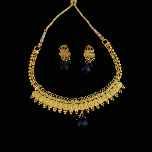 Gold-toned traditional necklace and matching owl-shaped earrings with dark blue teardrop beads, displayed on a dark gray reflective surface. The necklace has layered gold chains, coin-style discs, and dangling blue beads.