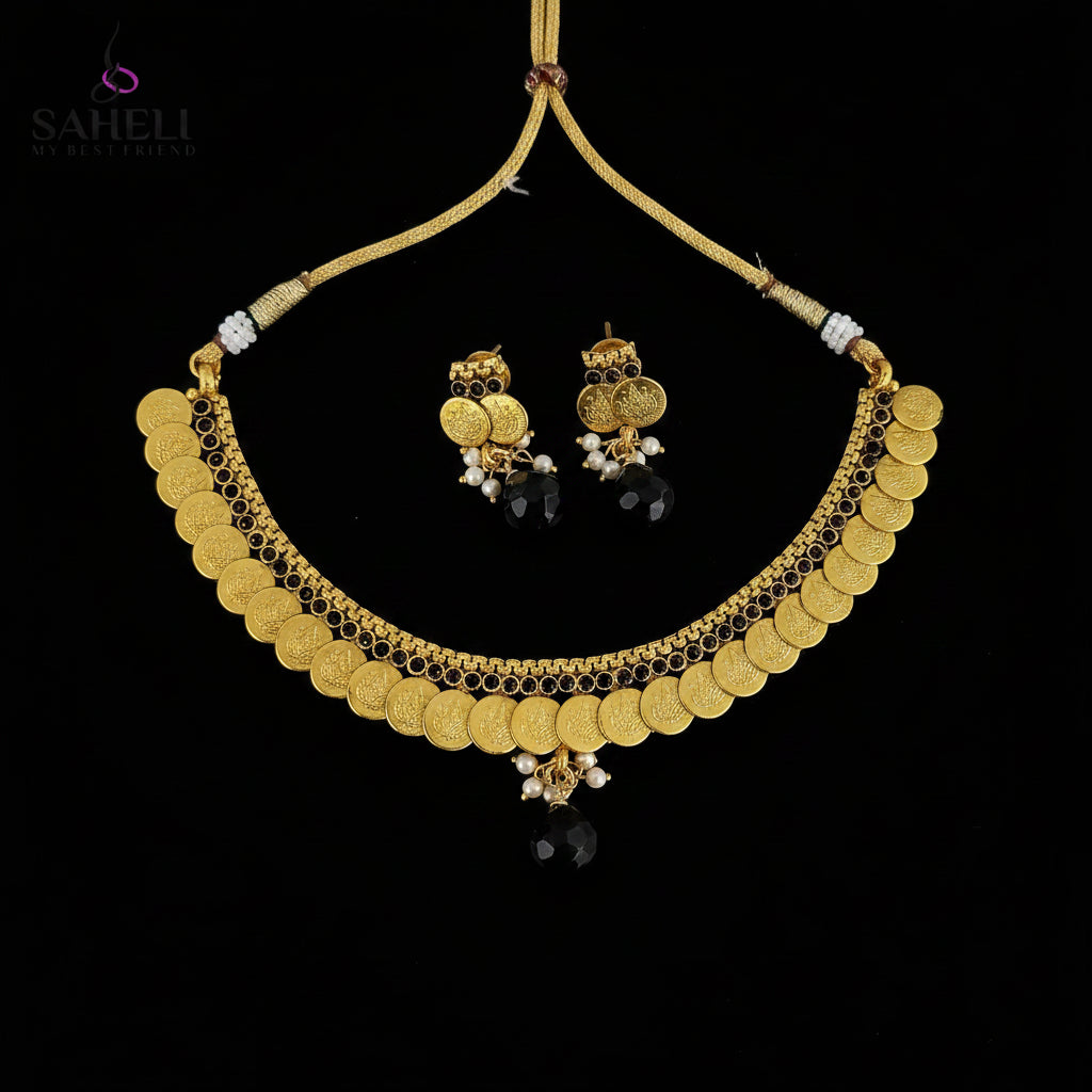 Gold coin necklace and matching earrings with black beads and white pearl accents, displayed on a dark reflective surface. The SAHeli by Saheli brand logo is visible in the top left corner.