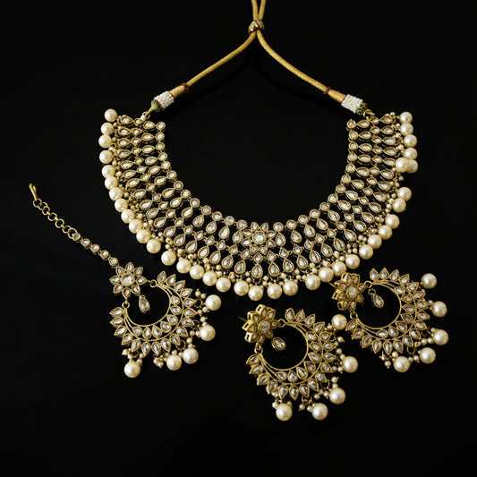 Gold-plated Indian traditional jewelry set featuring a choker necklace, matching earrings, and a maang tikka, embellished with faux pearls and clear stones, displayed on a dark reflective surface with the brand name 'SAHELI' visible.