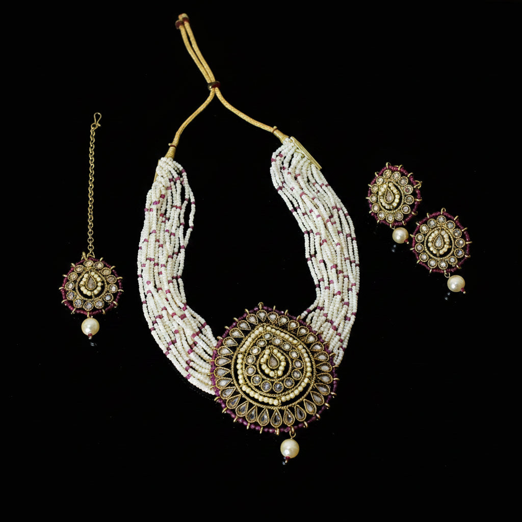 An ornate traditional Indian jewelry set featuring a multi-strand beaded necklace, matching earrings, and a maang tikka, displayed on a dark reflective surface. The jewelry is primarily composed of white beads with gold-plated filigree details and small red accents, with pearl-like white beads hanging at the ends.