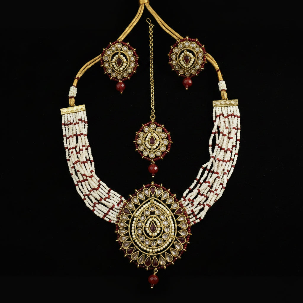 Gold and white beaded traditional Indian jewelry set featuring a necklace, earrings, and a maang tikka, displayed on a dark gray background. The set includes intricate gold-plated details, white seed beads, and red stone accents.