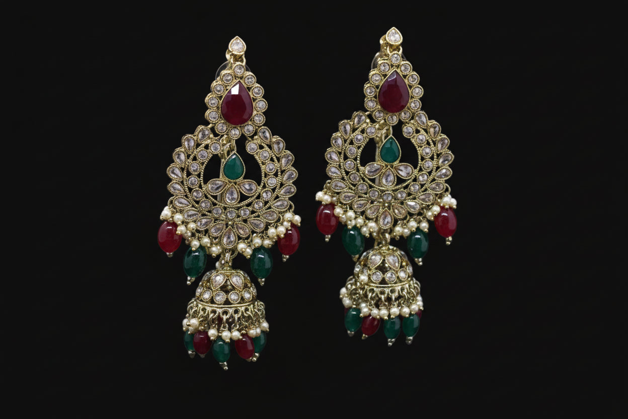 Ornate gold earrings with red and green stones on a stand, branded 'Saheli'.